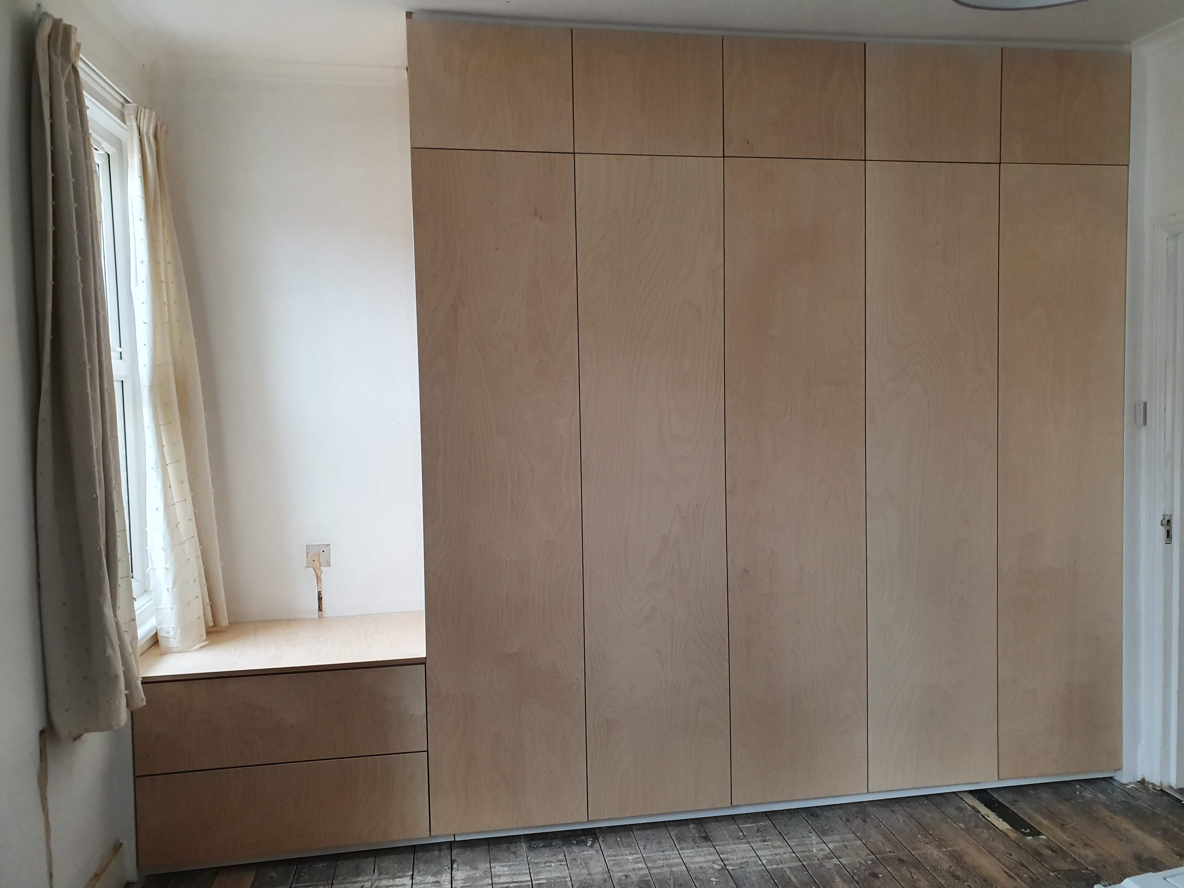 Minimalist fitted wardrobe in natural plywood with push-to-open doors, located in a bedroom.