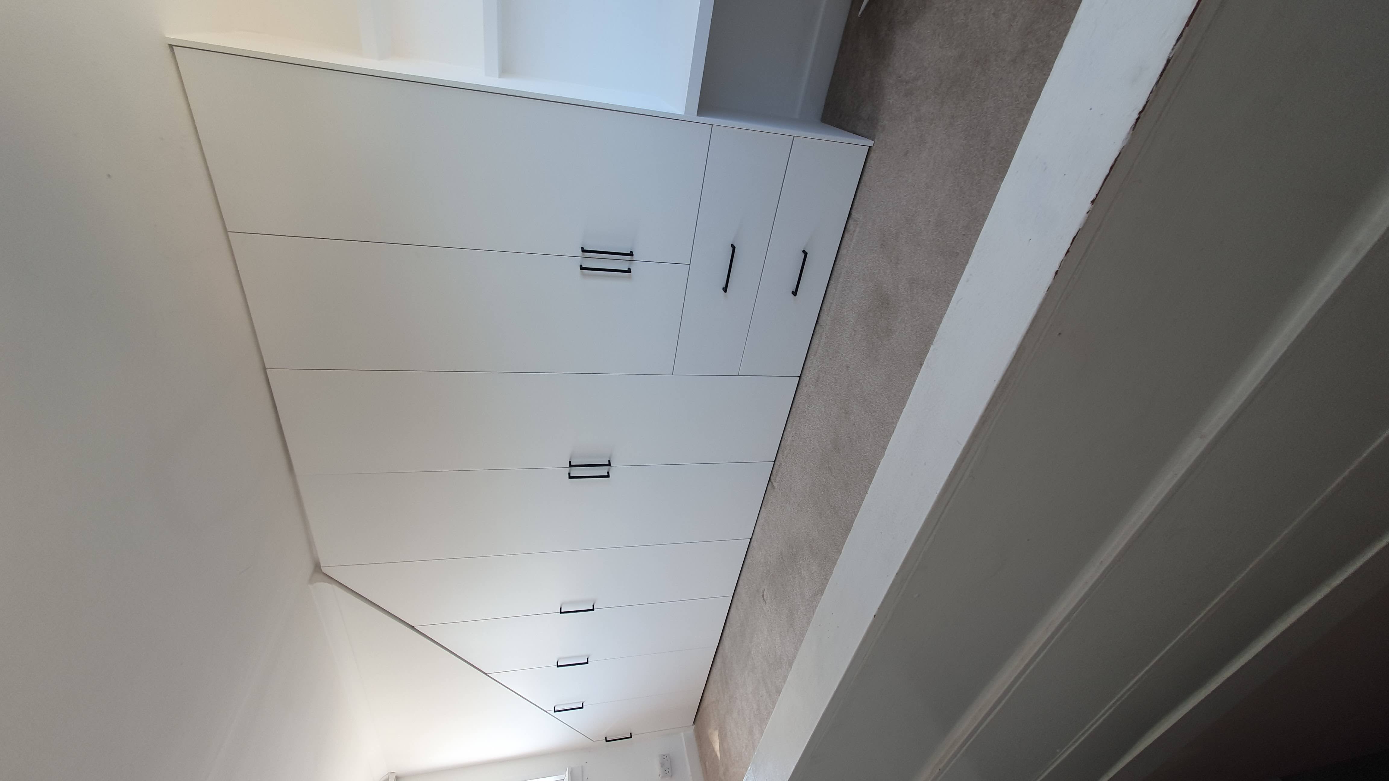 Modern white fitted wardrobes with black bar handles and an integrated desk, custom-built for a sloped ceiling bedroom.
