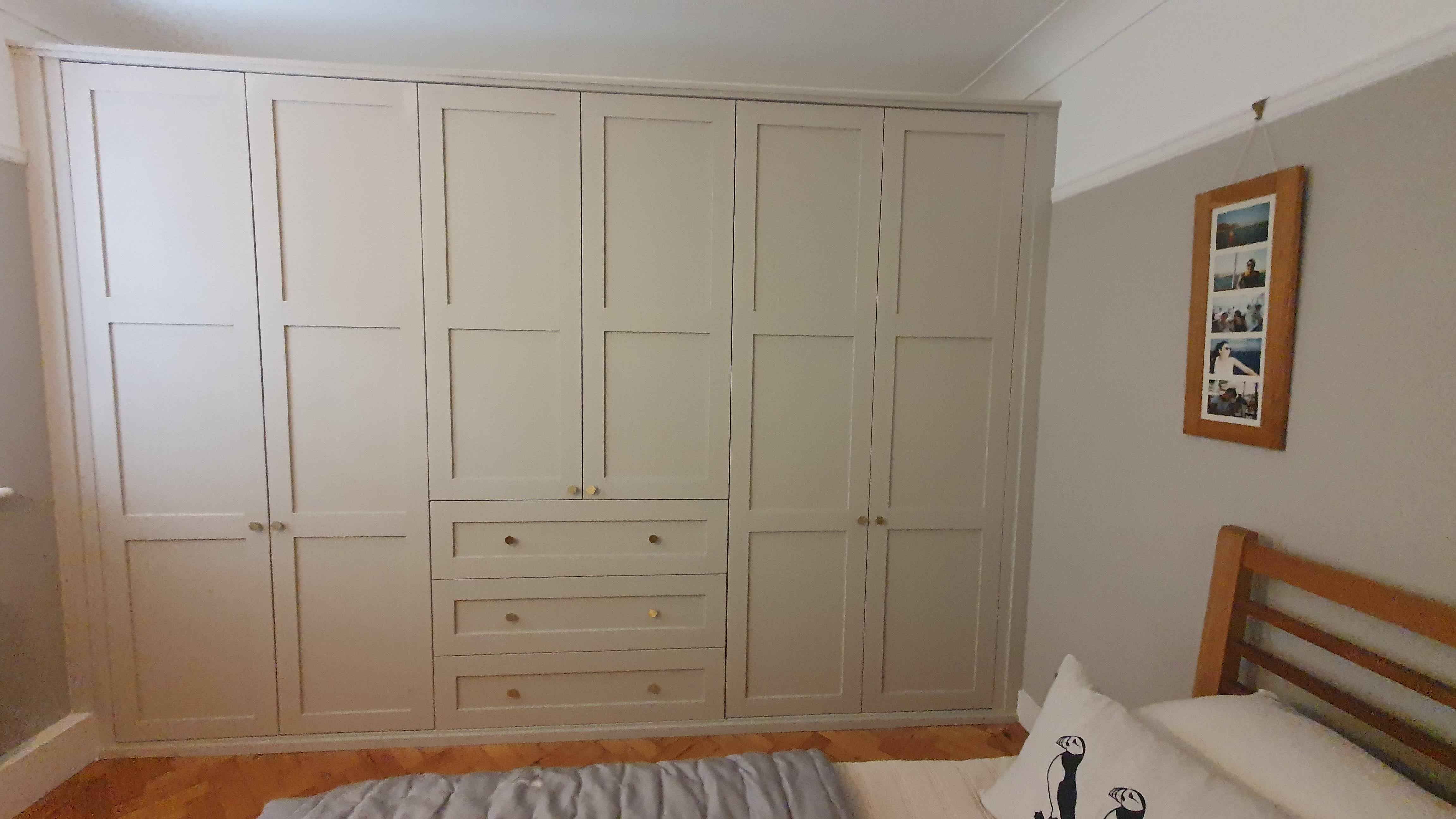 Fitted wardrobe in Farrow and Ball Skimming Stone with brass knobs, offering ample storage in a modern shaker style bedroom.