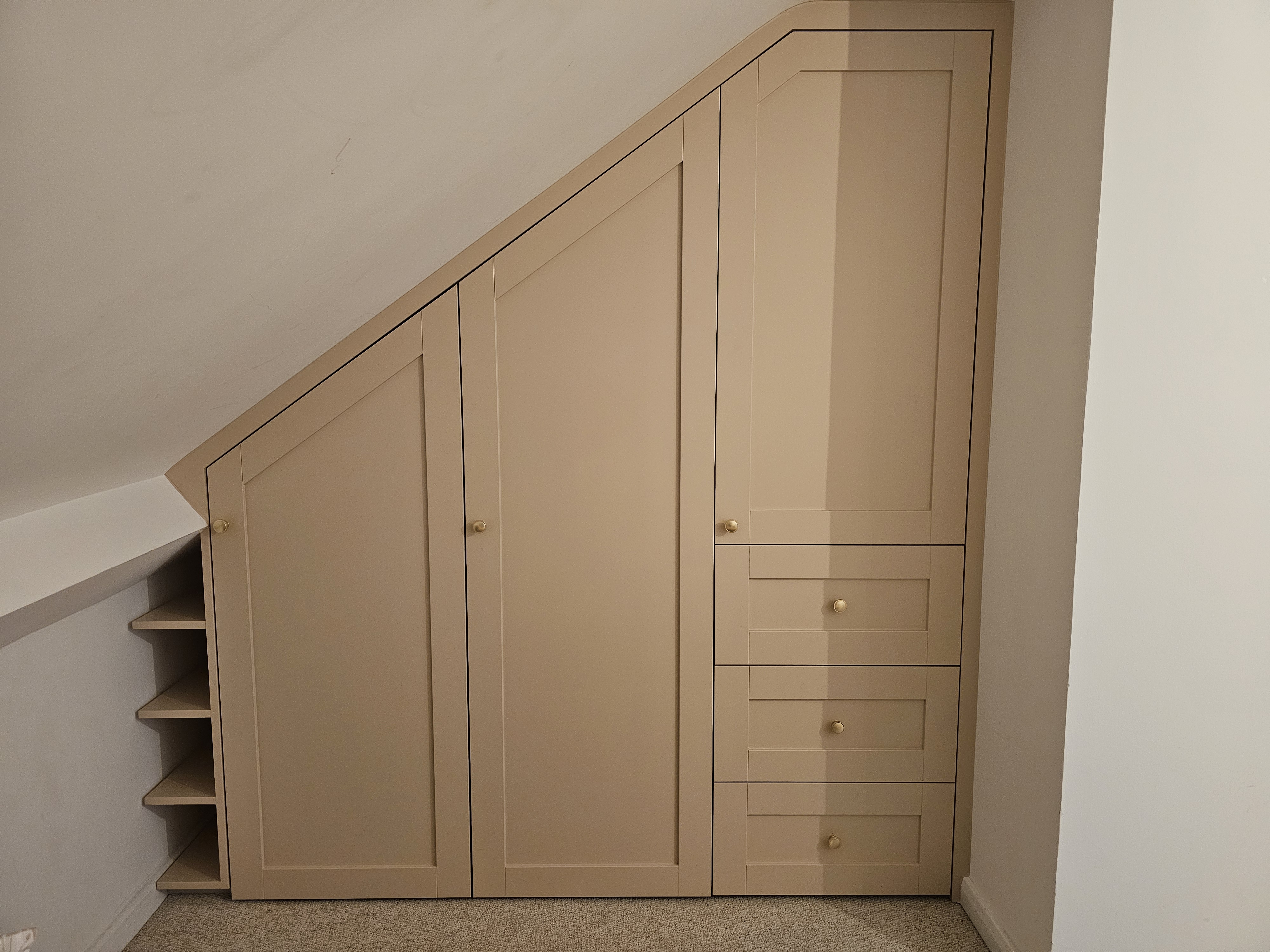 Fitted Shaker style wardrobe in Skimming Stone paint, designed for a sloped ceiling bedroom with brass knobs. Features integrated drawers and an open shelving unit.