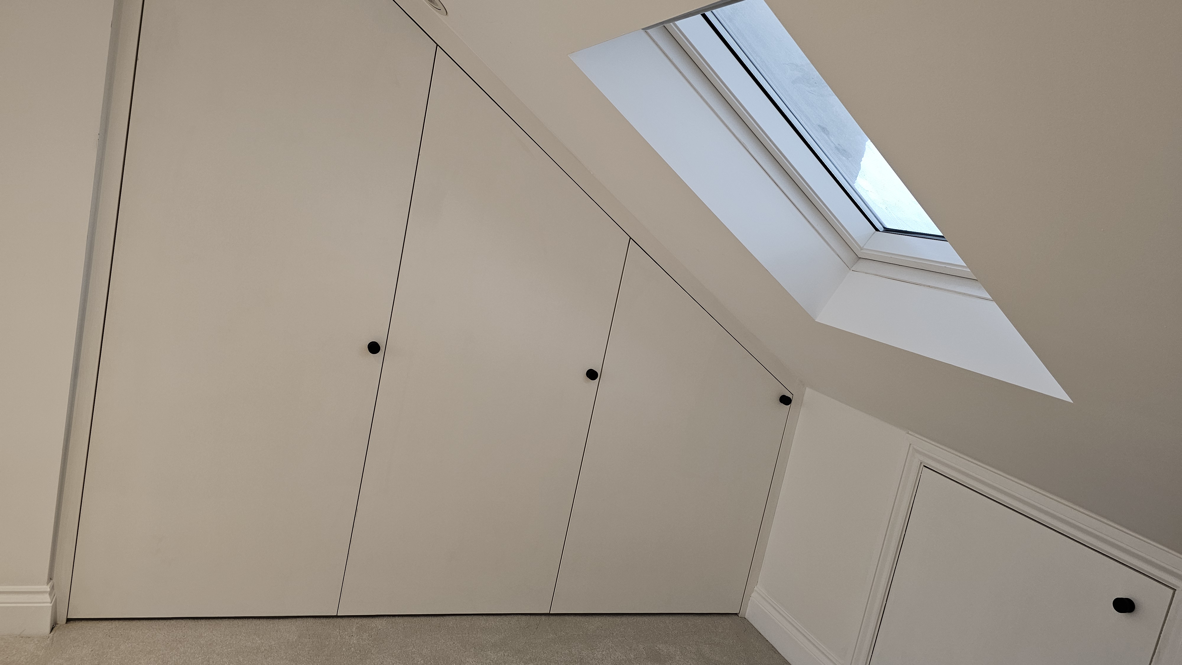 Minimalist fitted wardrobe in Dulux Timeless white, designed for a sloped ceiling attic bedroom, featuring black knobs.